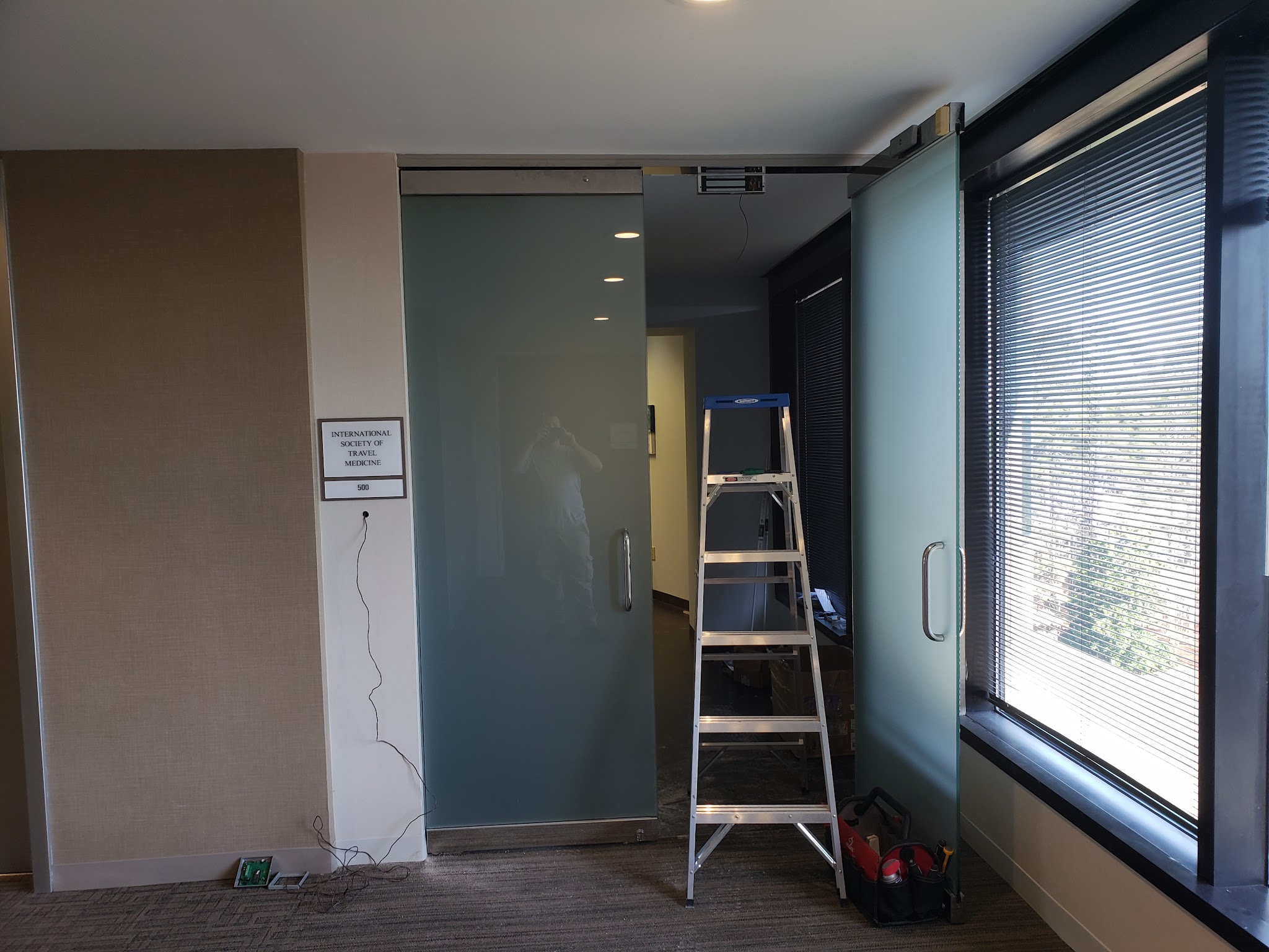 Glass office door installation in progress with a ladder and tool bag